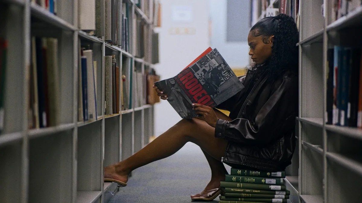 Jeune femme assise sur une pile de livres entre deux rayonnages de bibliothèque, plongée dans un grand livre illustré