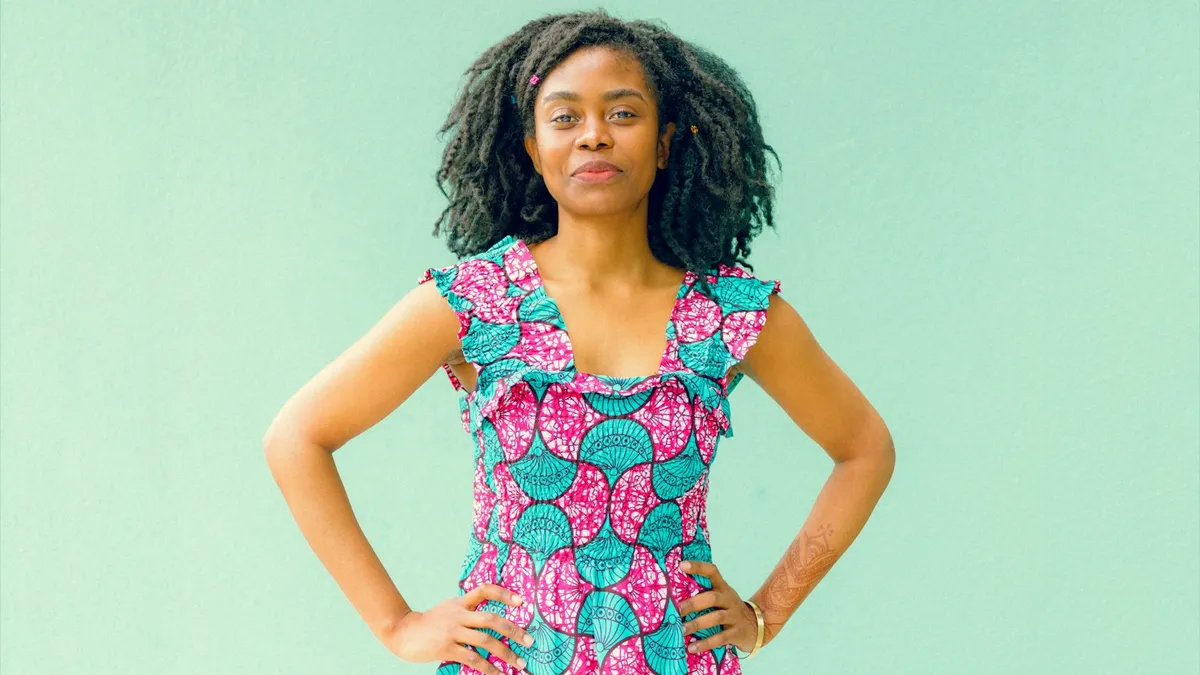 Femme souriante vêtue d'une robe en wax rose et turquoise, mains sur les hanches, devant un mur vert d'eau
