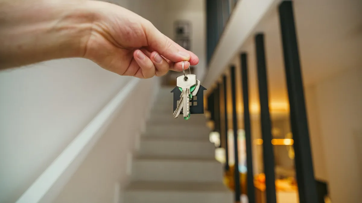 Main tenant un trousseau de clés avec un porte-clés en forme de maison, devant un escalier