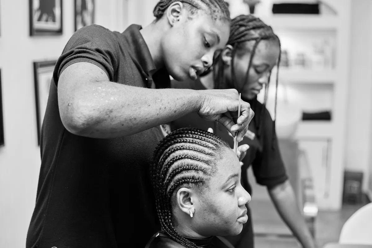 Coiffeuse réalisant des nattes collées sur une cliente dans un salon, en noir et blanc